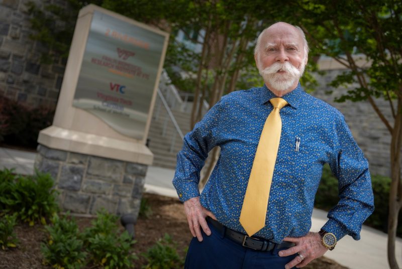 Man with blue shirt and yellow necktie standing with hands on hips outside with VTC sign in background