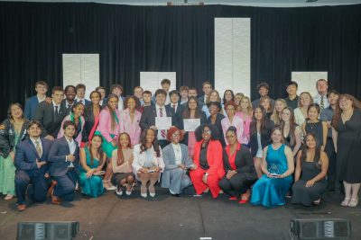 A large group of smiling individuals poses together on a stage, dressed in business and ballroom attire. Some individuals hold paper certificates.