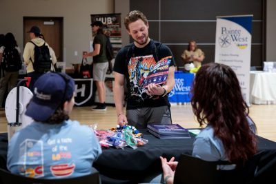 Two people sit at a table in the foreground, facing away from the camera. A student in jeans and a graphic t-shirt and wearing a backpack examines a full-color flyer on the other side of the table. In the background are more tables with names and logos of apartment management companies and students browsing among them.