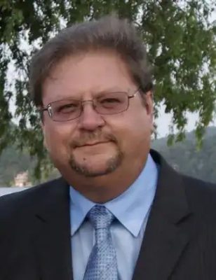 Dennis Hall smiles while wearing a suite and tie and standing in front of green trees.